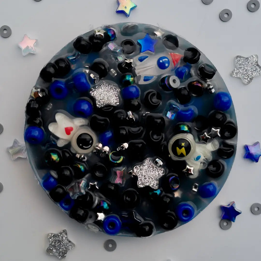 Treasure Cove Pick-Me Pad – overhead view of dark galaxy-themed sensory pad with black and blue beads, silver stars, and character charms